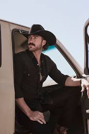A man sitting in his truck wearing black clothes with a cowboy hat.